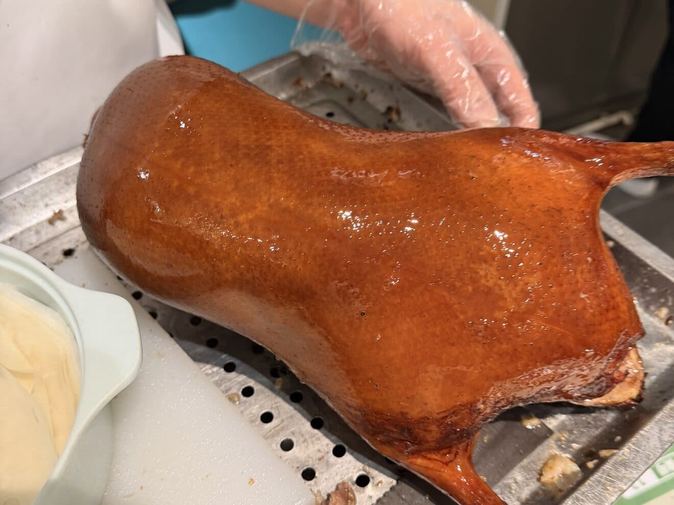 A whole roast duck on a board: glazed skin and leg, a gloved hand carving slices with a knife