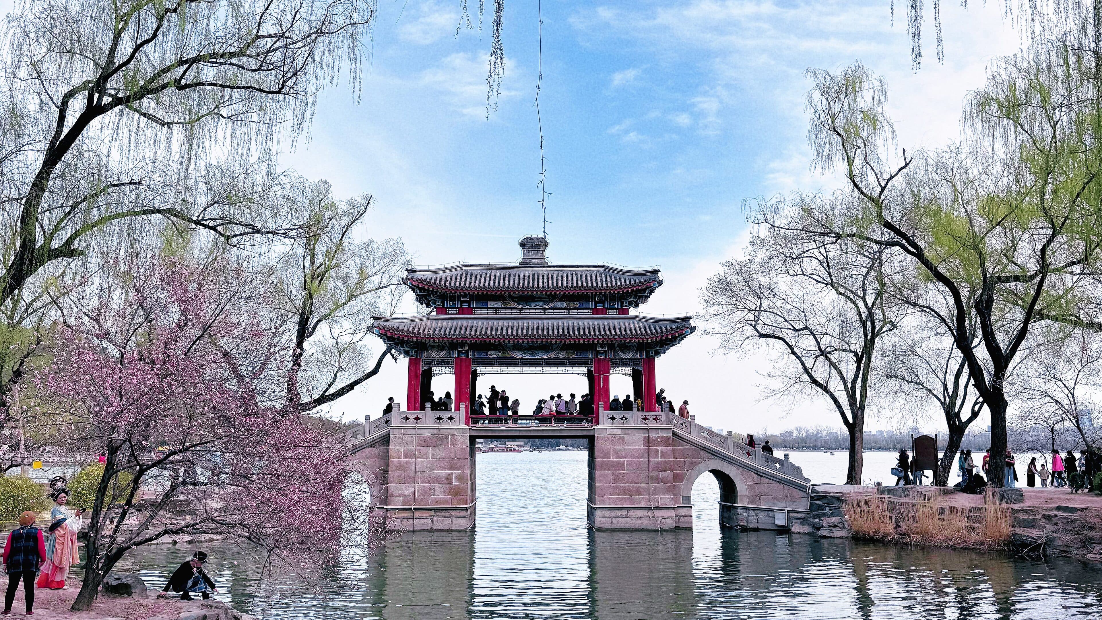 A red-columned pavilion bridge on a stone arch, the lake below, and willows and pink blossom on the bank