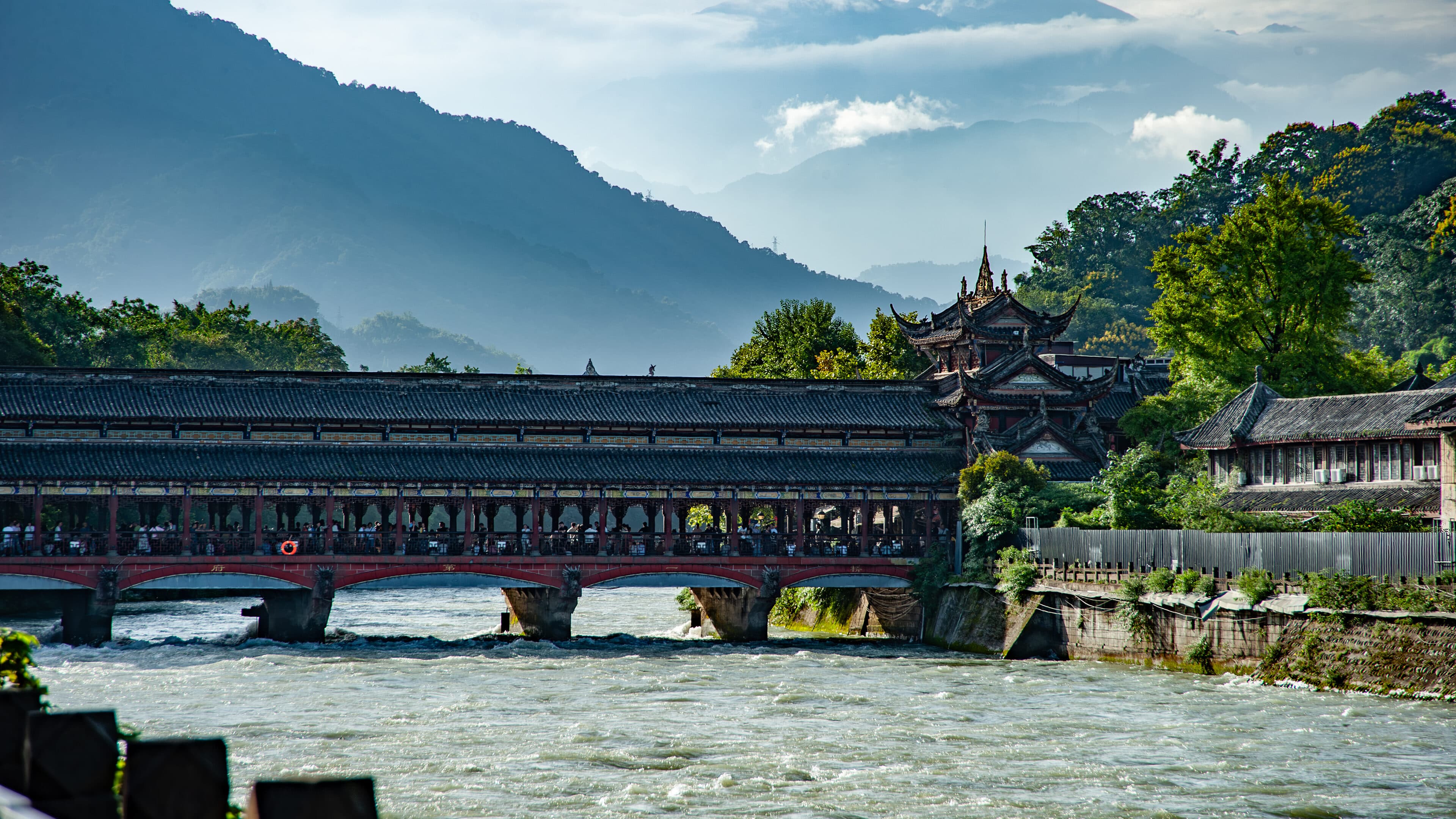 A covered timber bridge across the river, with traditional eaves and green hills on both banks