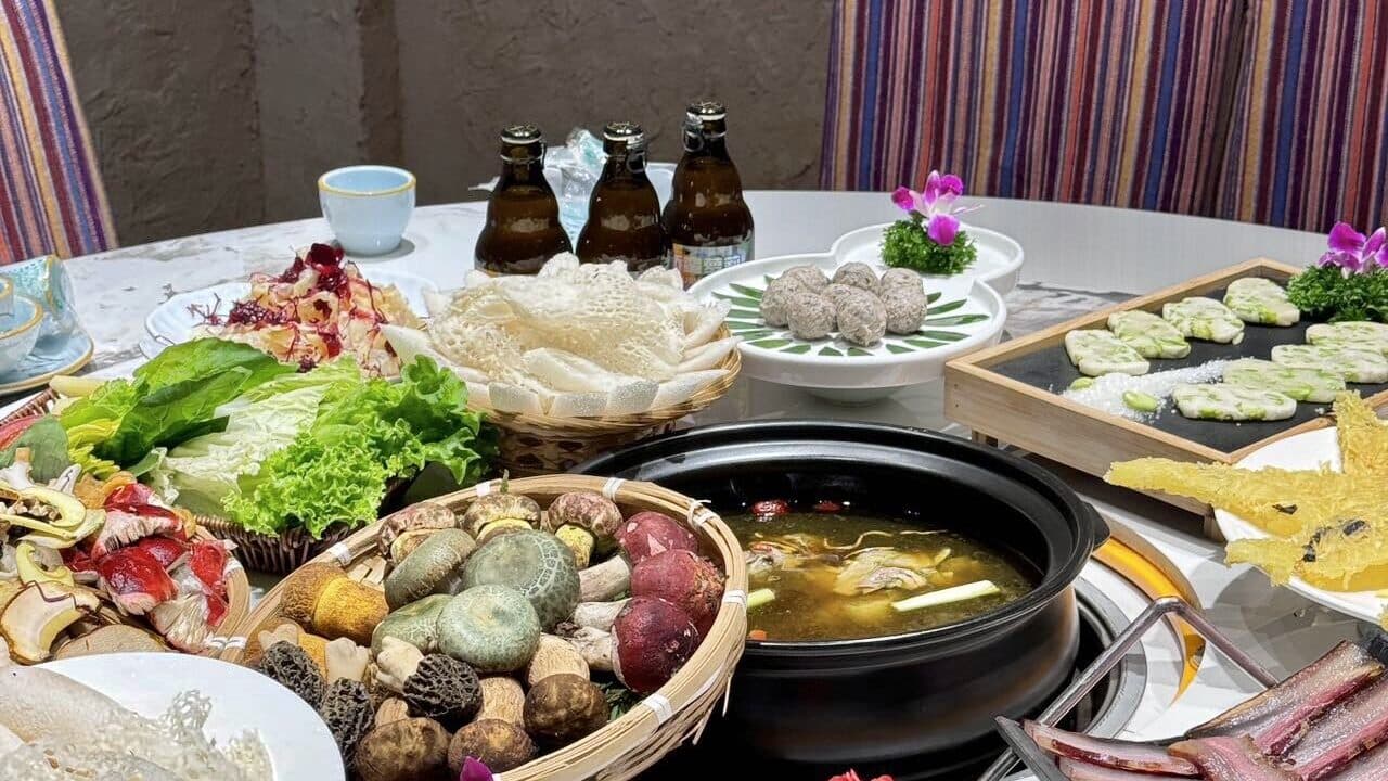 A round home table with a clay-pot soup, a basket of mushrooms and plates of dishes, striped chairs behind