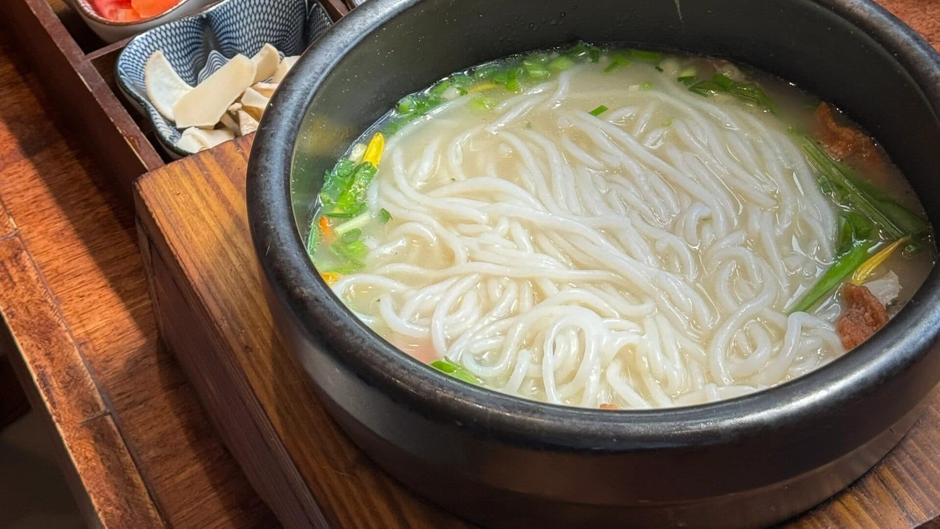 A stone pot of Yunnan rice noodles in broth beside a wooden tray of small side dishes