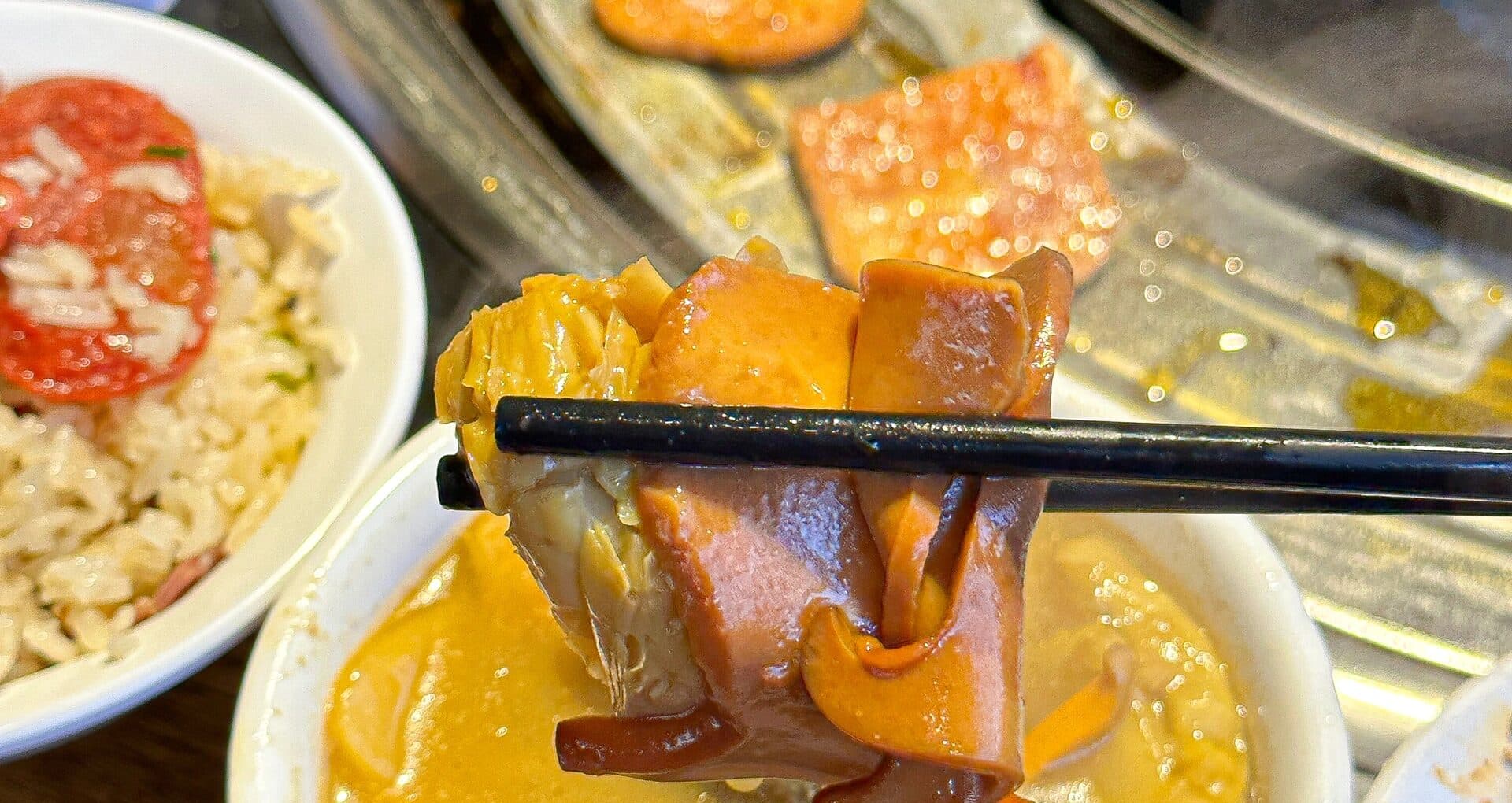 Chopsticks lifting a piece from a golden broth pot, with rice and side dishes alongside