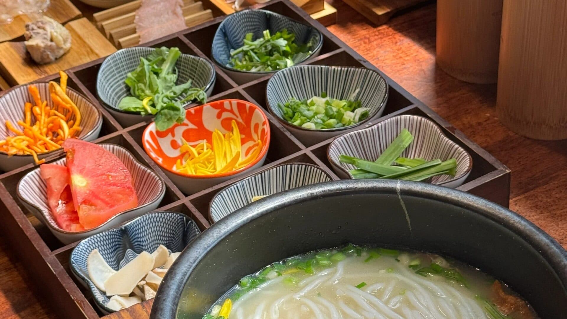 Crossing-the-bridge noodles: small side dishes in a wooden tray beside a bowl of rice noodles in broth with spring onion