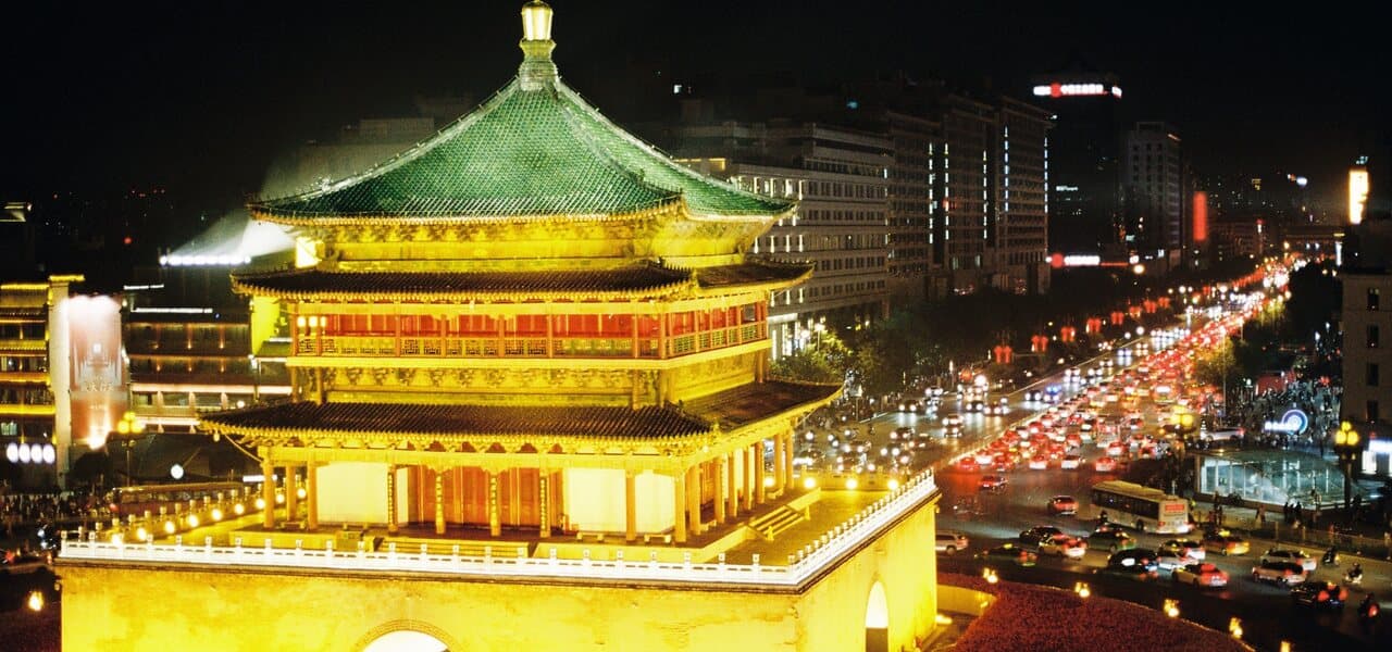 Xi'an's Bell Tower lit at night with green glazed-tile roofs above its brick base, car light trails on the roundabout beside it