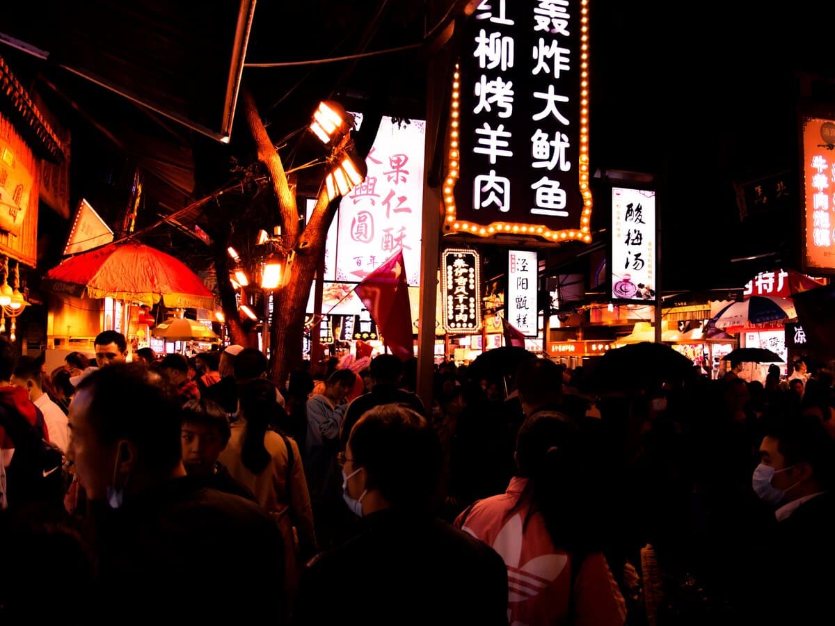 A night food street with wet reflective paving, rows of vertical lightbox signs and passers-by in backlit silhouette