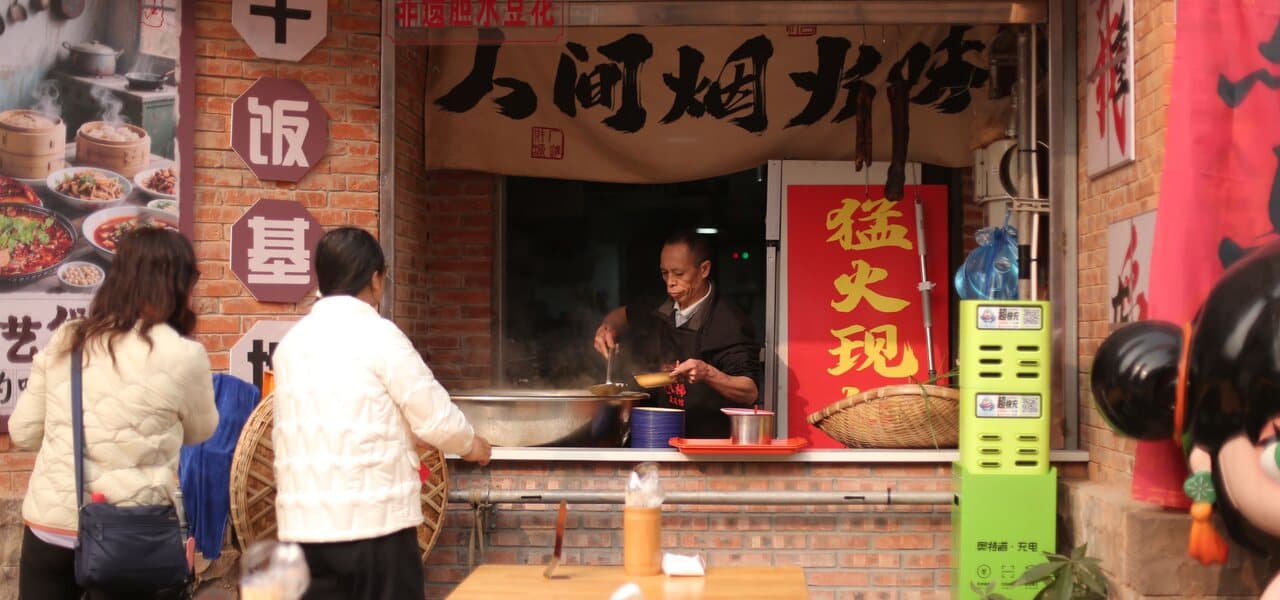 A street food stall: a cook working at a griddle with steam rising inside a brick-framed window, two customers standing with their backs to the camera