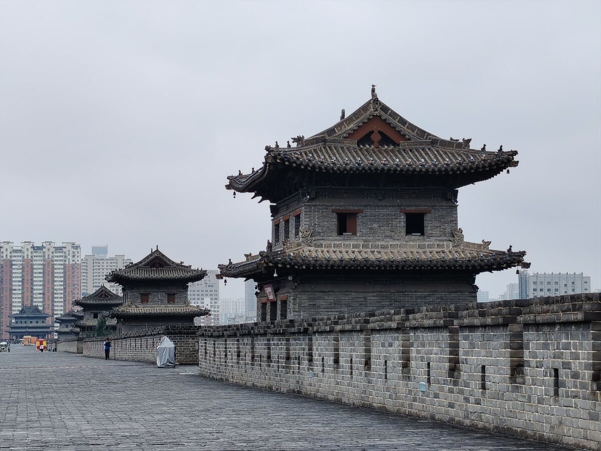 Xi'an's city wall in daylight: the brick walkway runs toward a watchtower, with modern blocks under an overcast sky beyond