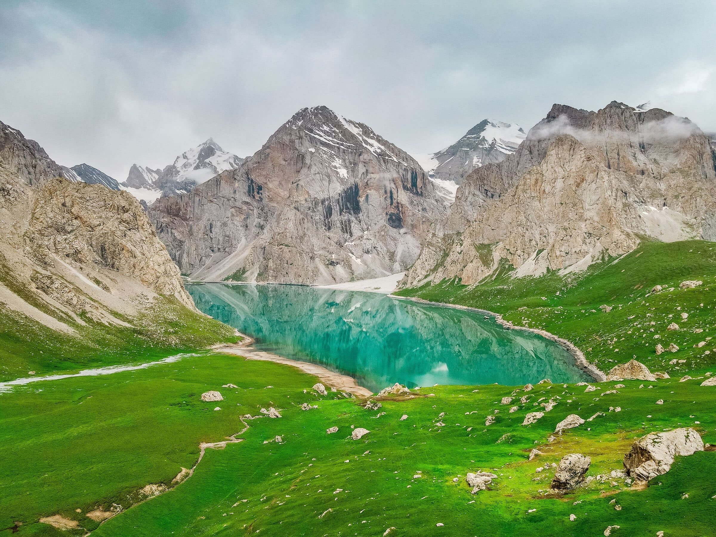 A turquoise alpine lake below snow-capped Tianshan peaks, with a trail crossing the meadow