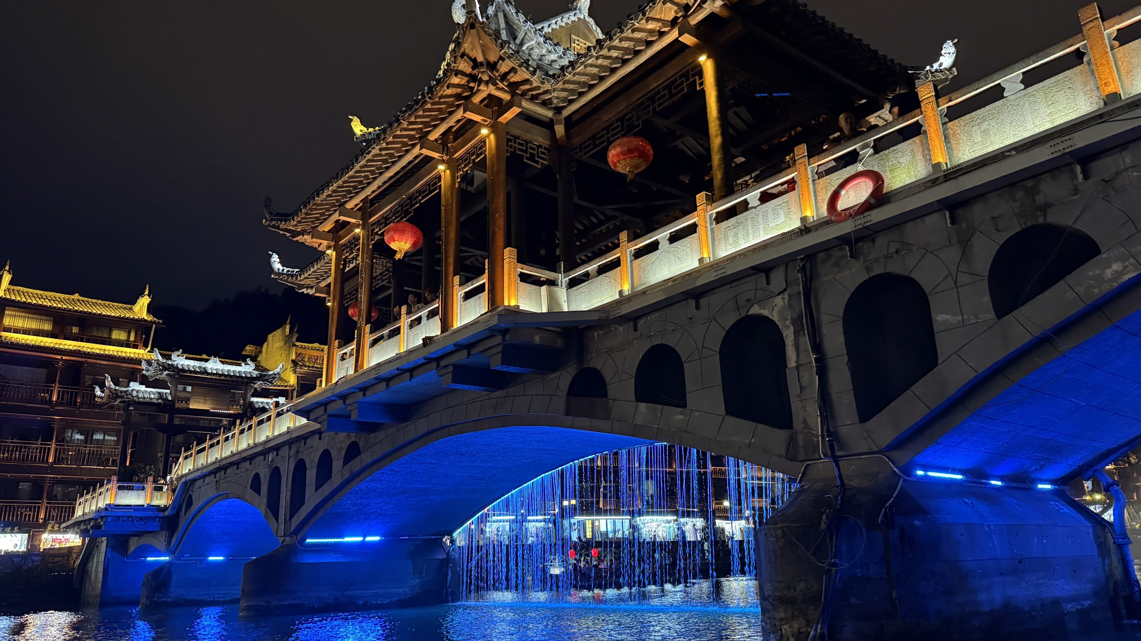 A multi-arch stone bridge with a covered gallery at night, red lanterns on the balustrade and a blue-lit water curtain between the arches