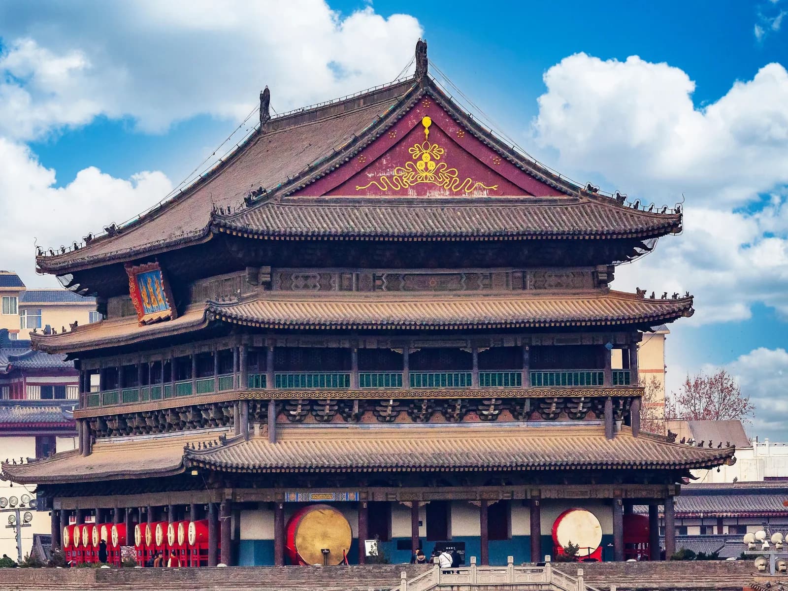 Xi'an Drum Tower in daylight under a blue sky: a triple-eaved pavilion with green glazed-tile roofs and a row of large red drums beneath the eaves, above its brick base and stone-railed steps