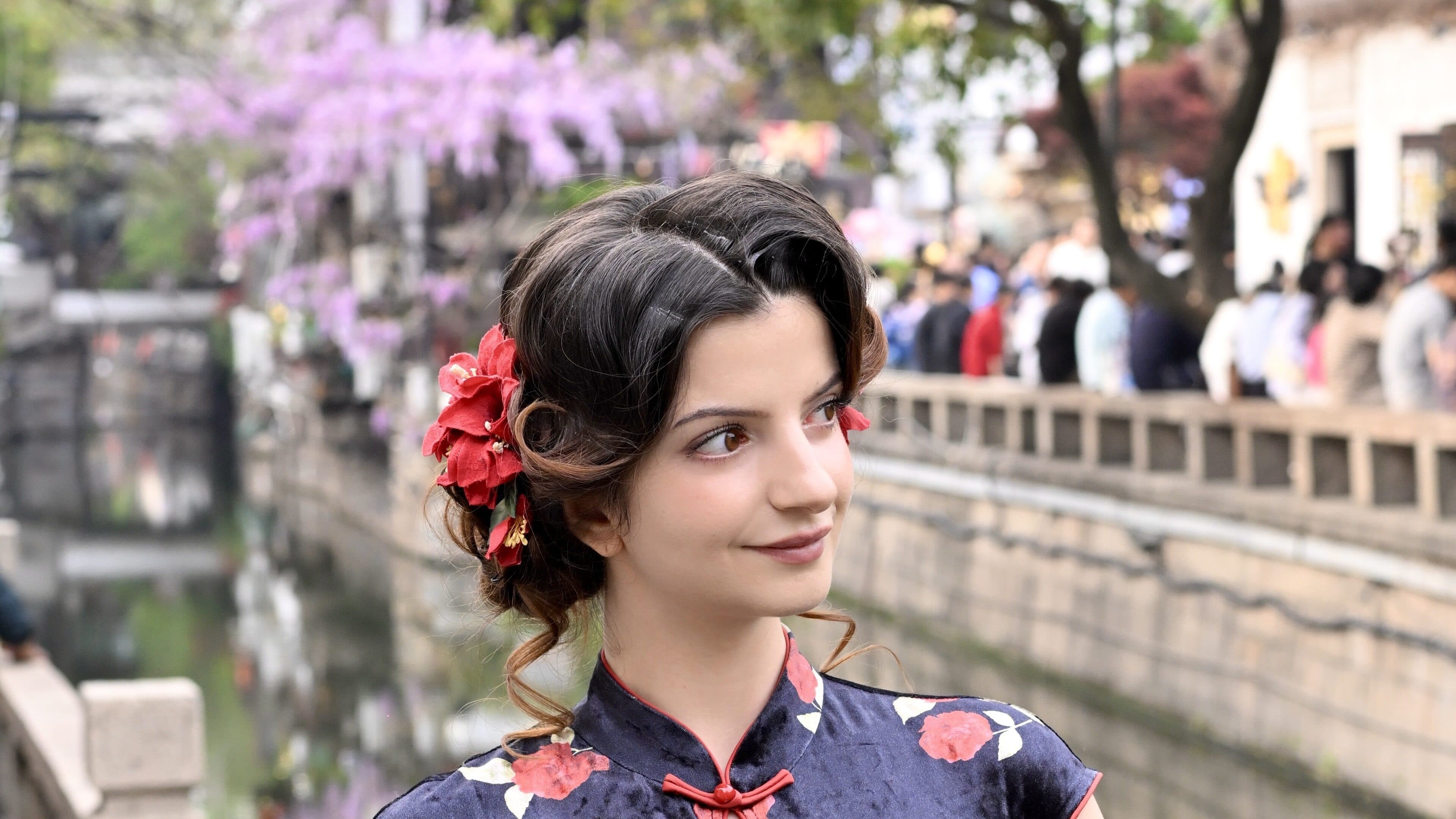 A woman in a dark blue floral qipao on a canal-side stone bank, with a stone bridge and visitors behind