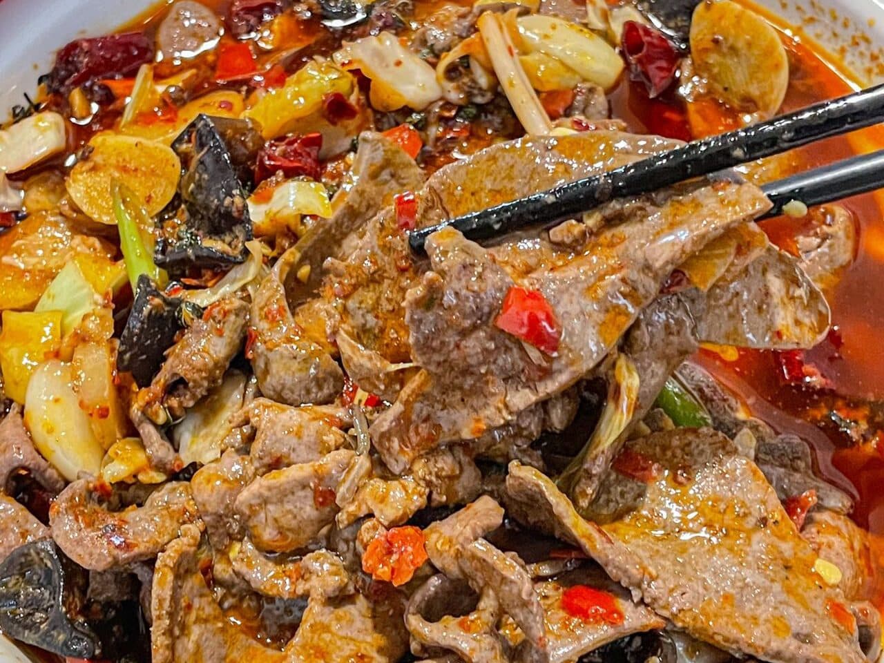 Sliced meat and vegetables in red chilli oil, a piece lifted with chopsticks