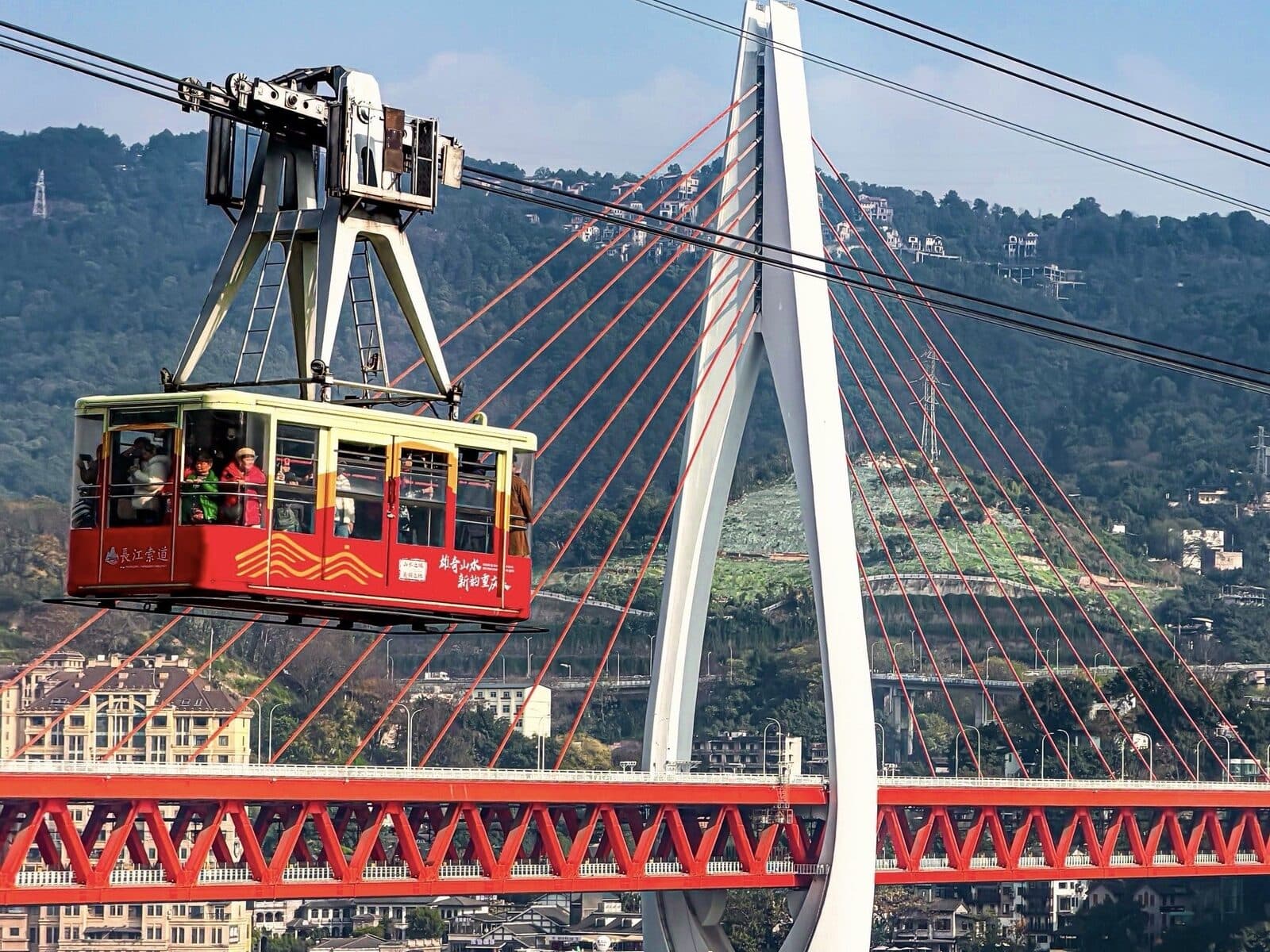 The Yangtze River cableway in Chongqing by day: a car hanging above the river with the Dongshuimen bridge and the water below