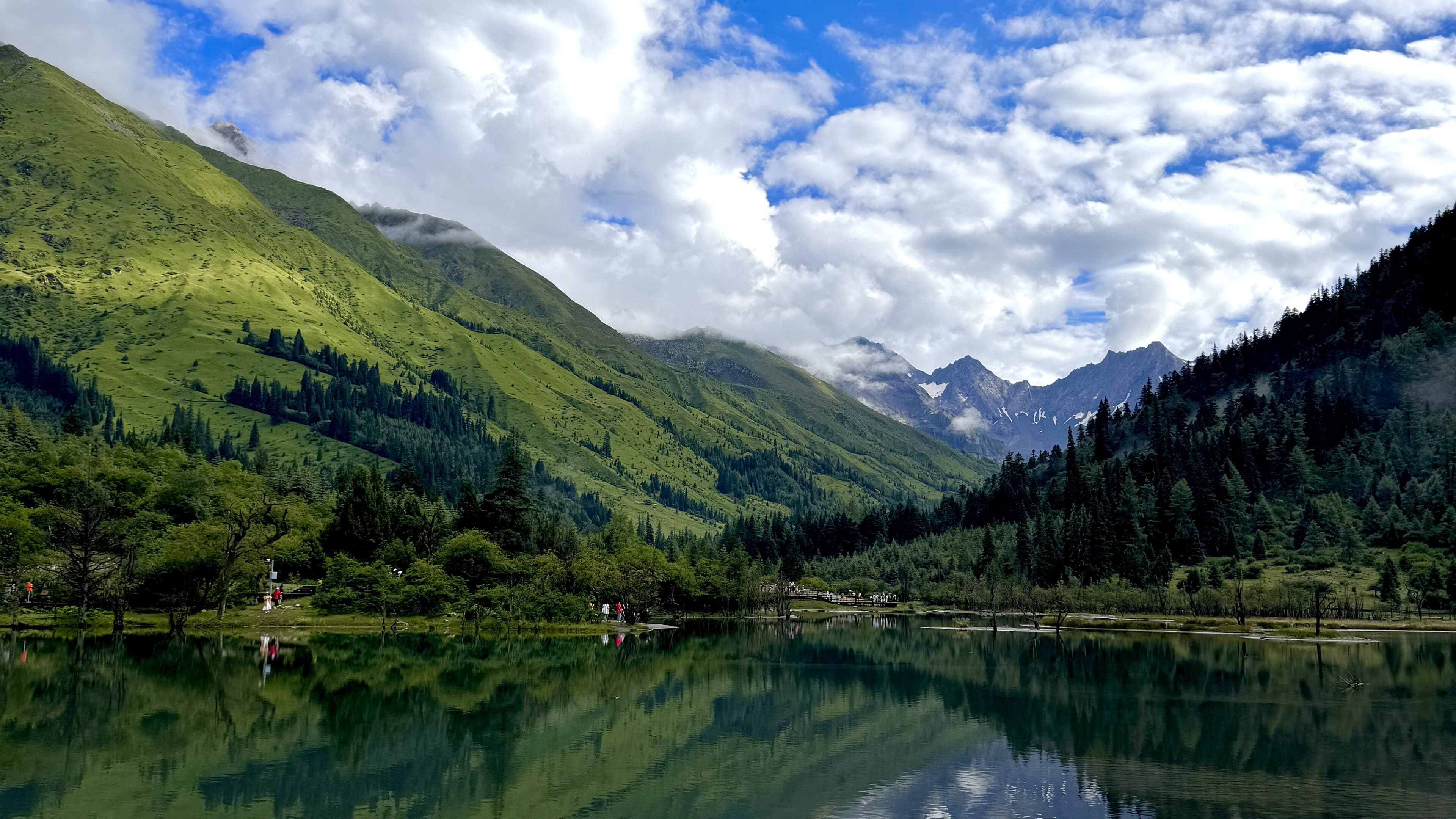 Alpine meadow and a mirror-still lake, rocky snow peaks and cumulus clouds beyond