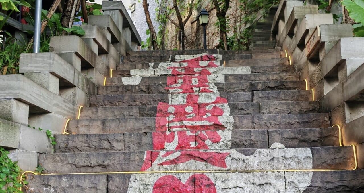 A hillside stairway climbing between stone retaining walls, greenery and lamps