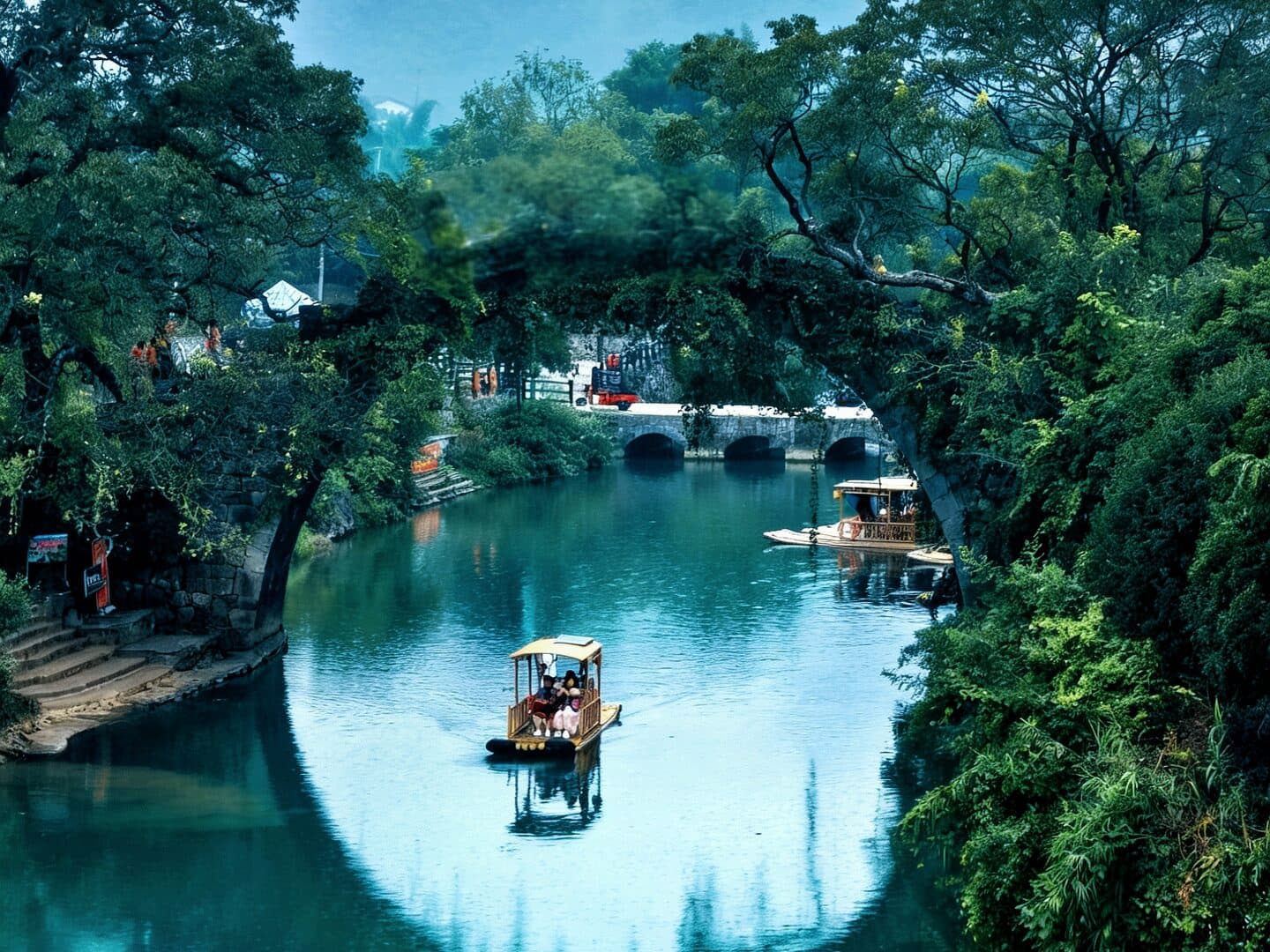 A green river and stone arch bridge between karst hills, bamboo on both banks and peaks beyond, with a bamboo raft on the water
