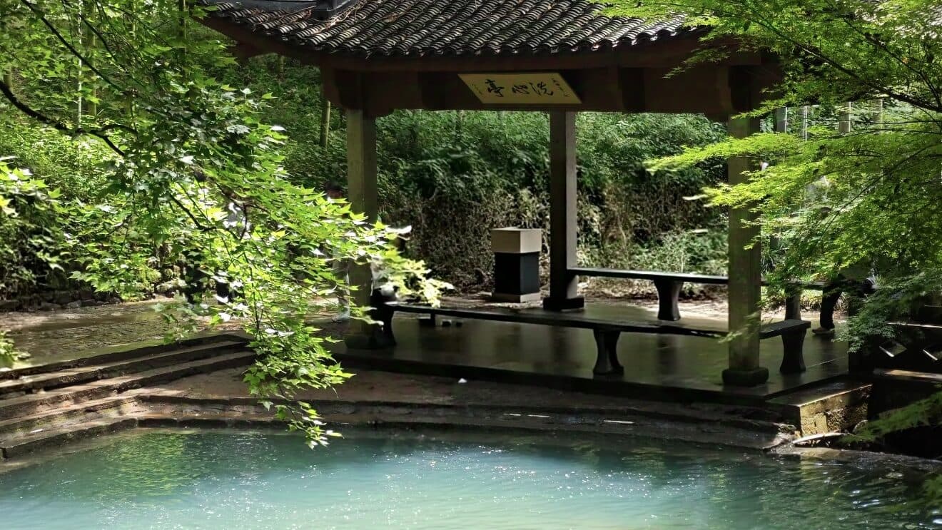 A timber pavilion beside a pond, with benches inside and green trees reflected in the water