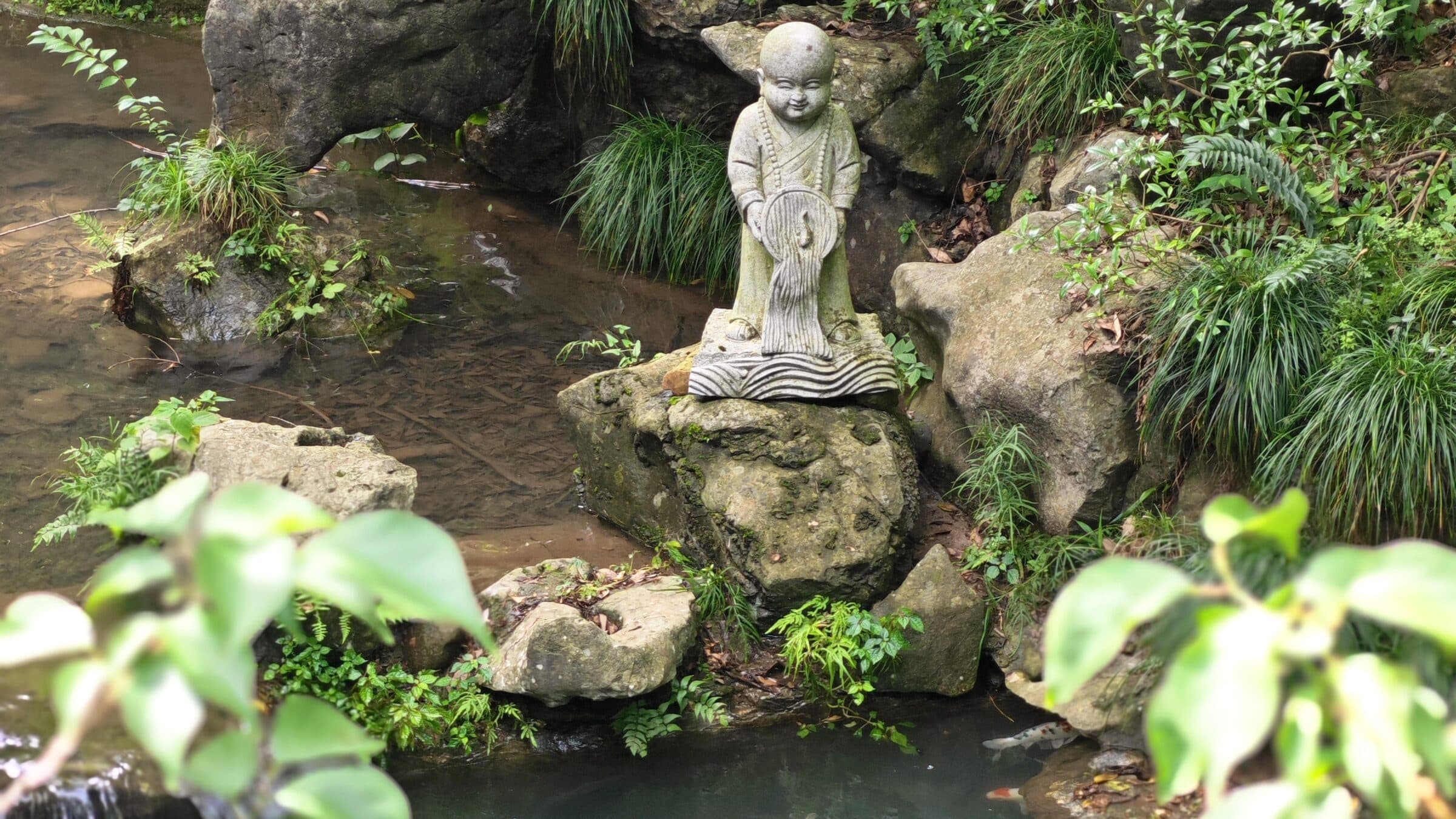 A stone-carved monk figure among mossy rocks, ferns and shallow water at Feilai Peak