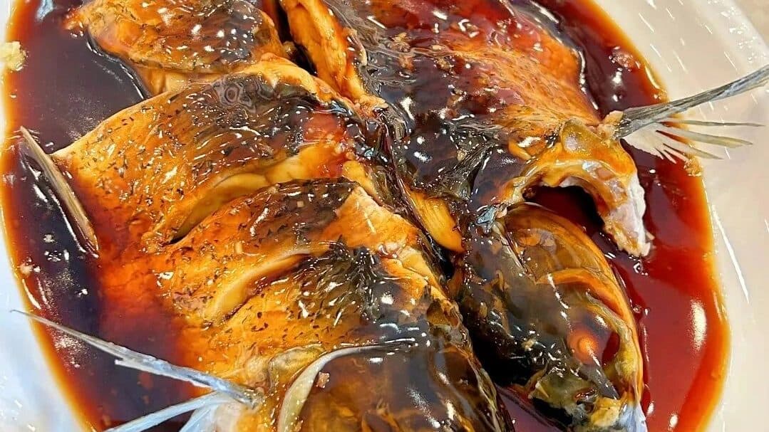 A whole braised fish in dark sauce on a white plate, scored and ready to share