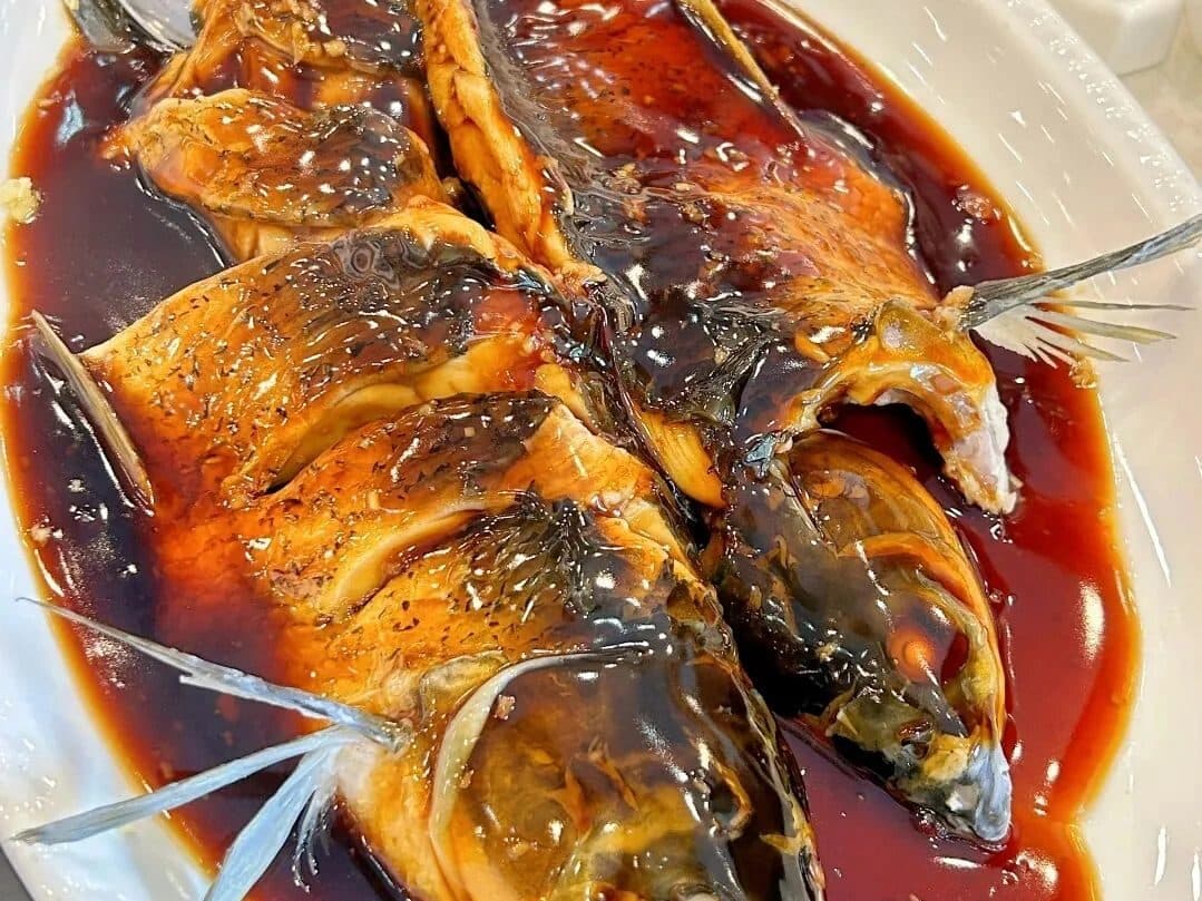 A whole braised fish in dark sauce on a white plate, scored and ready to share