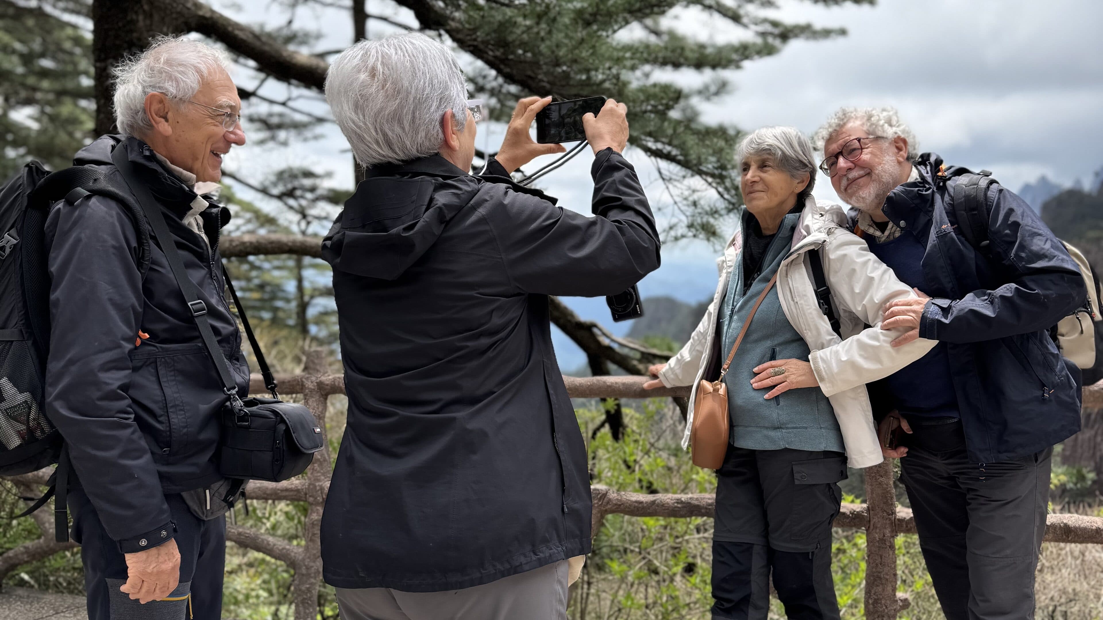 Three visitors at a mountain path railing, one raising a camera to photograph the others