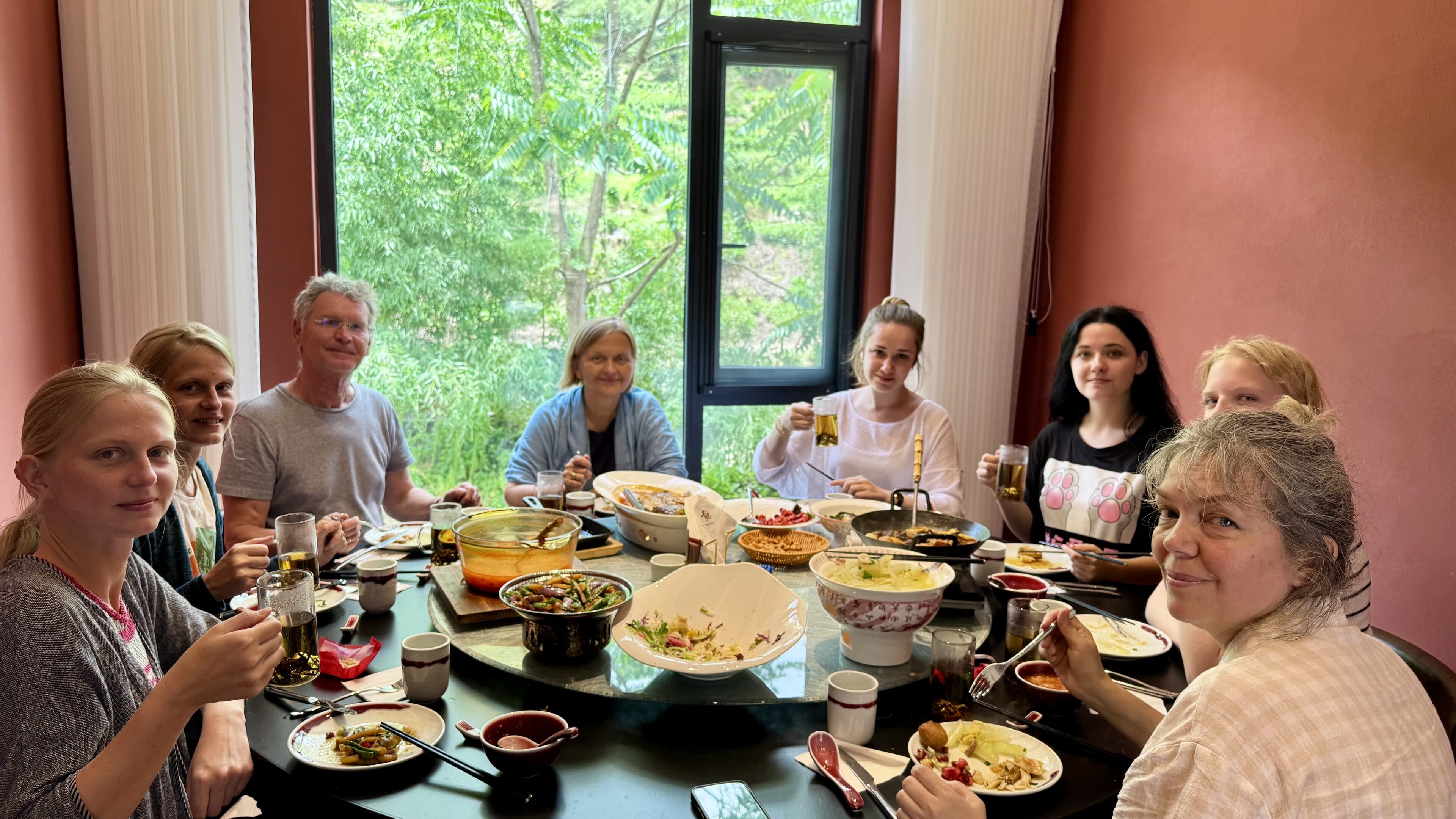 People around a round dining table heaped with dishes, green leaves beyond the window