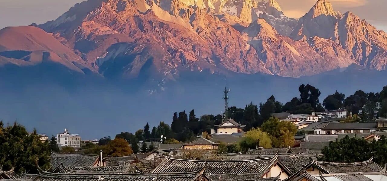Dusk over ranks of tiled rooftops and trees, with a distant snow-mountain ridge in warm light