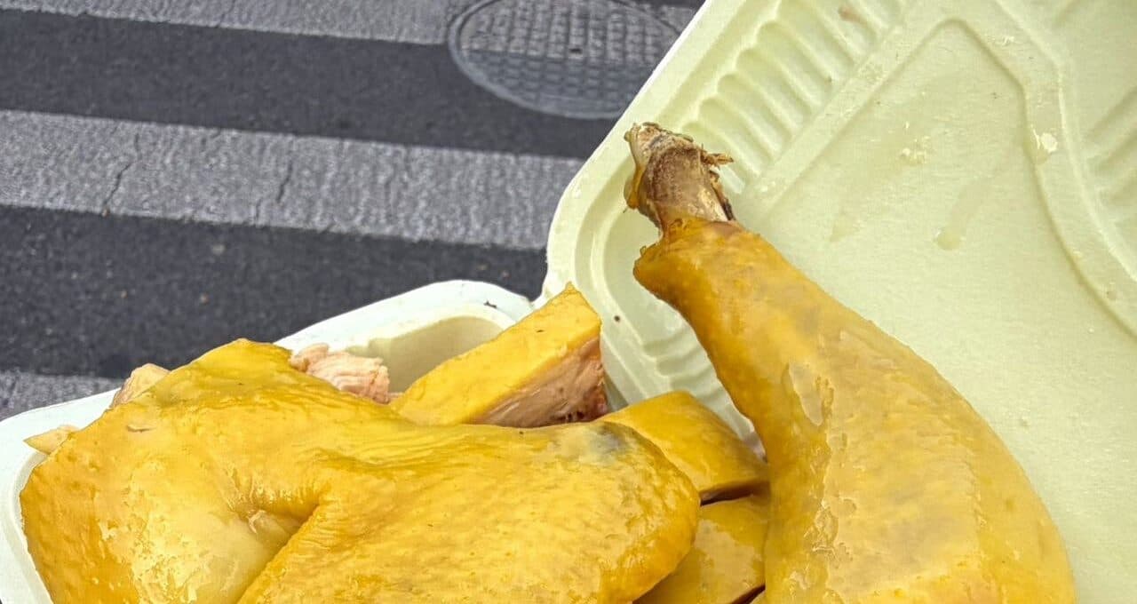 A whole yellow-skinned cooked chicken in an open takeaway foam box, with a street crossing and pavement behind