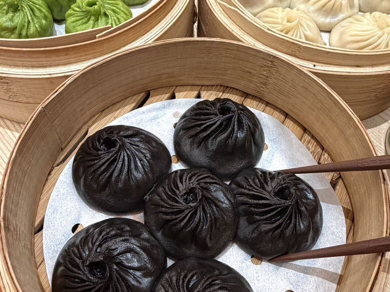 A three-tier bamboo steamer with green, white and black-truffle xiaolongbao, chopsticks lifting one