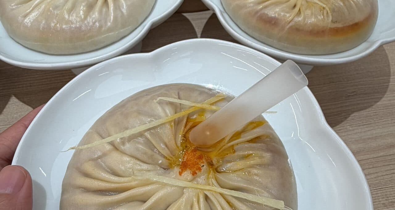 A soup-dumpling close-up: one large dumpling with a straw and ginger on top, two steamed buns in white dishes beside it