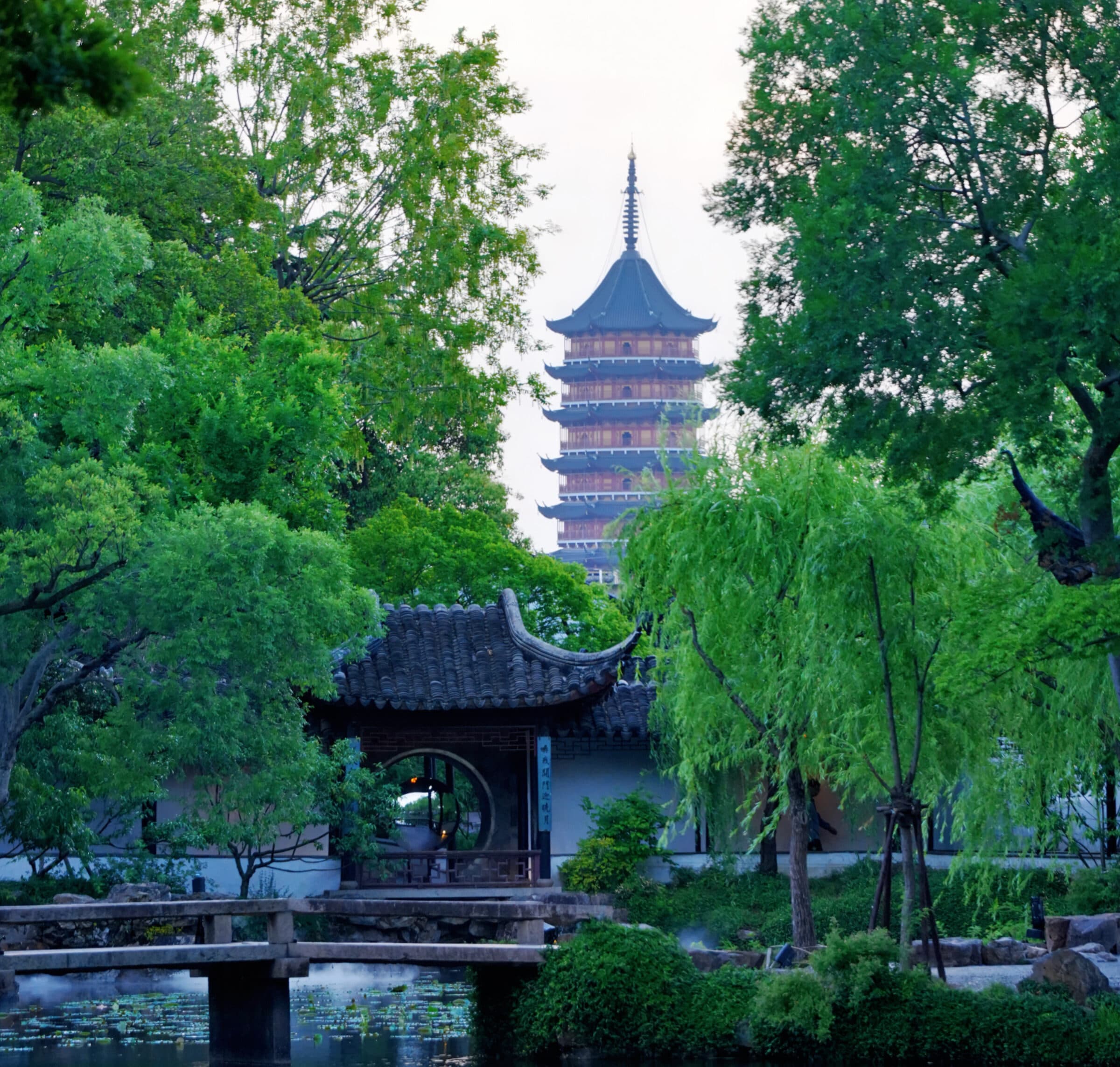 A Suzhou garden pond and zigzag bridge, willows framing the Tiger Hill pagoda beyond white walls and grey tiles along the water