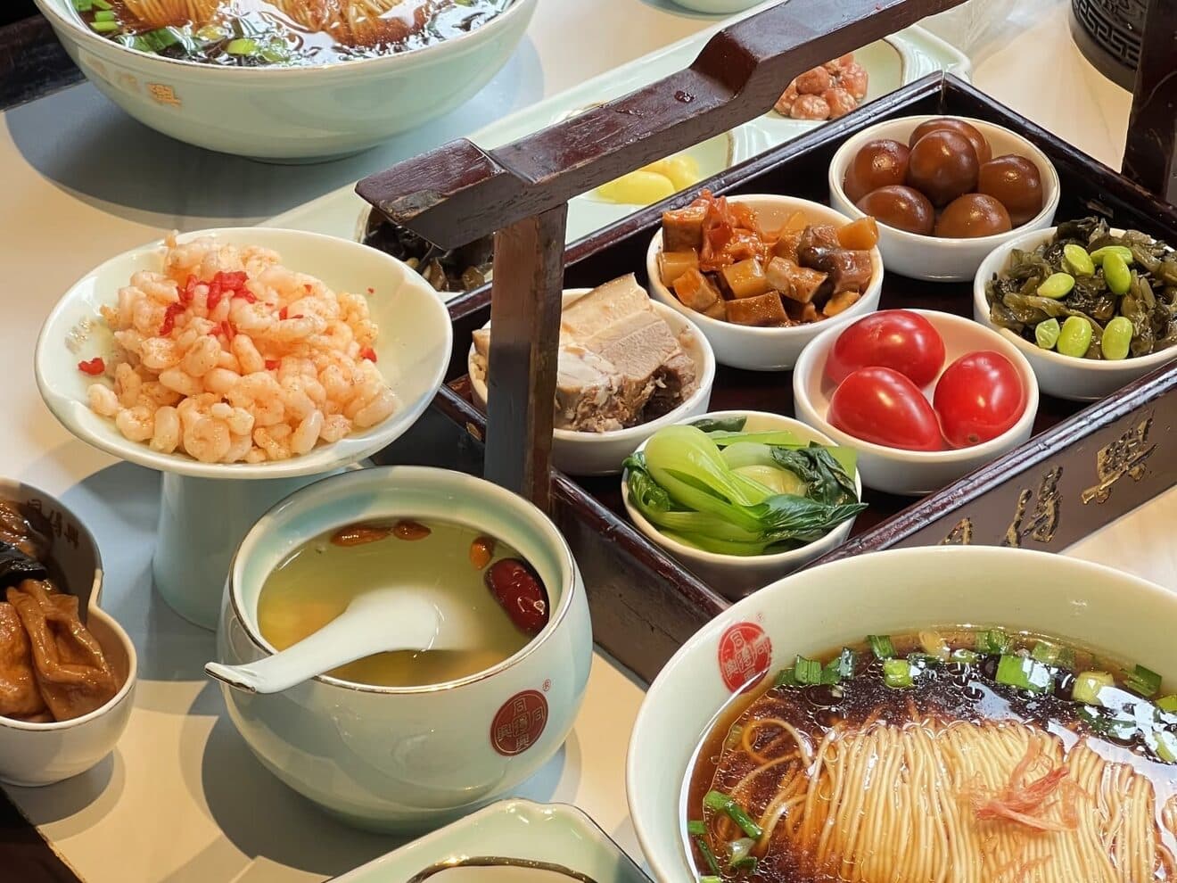 A bowl of fine Suzhou noodles in clear broth with greens, with small side dishes and a soup bowl beside it