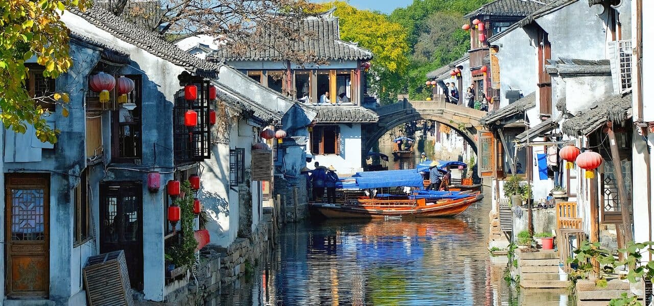 A water-town canal: white-walled, grey-tiled houses and red lanterns along the banks, a blue-canopied boat on the water, an arch bridge beyond, all reflected in the stream