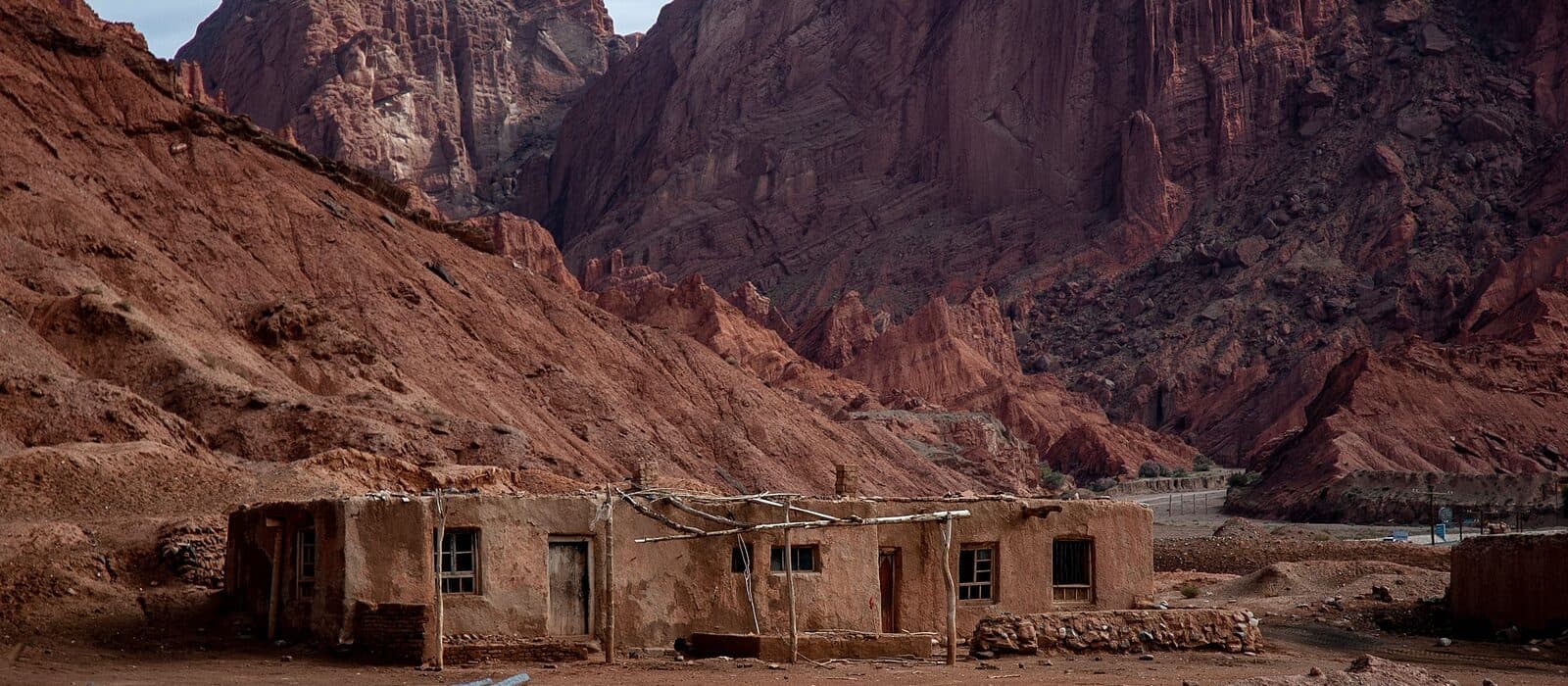 A weathered mud-brick dwelling with a timber pole frame below red rock cliffs