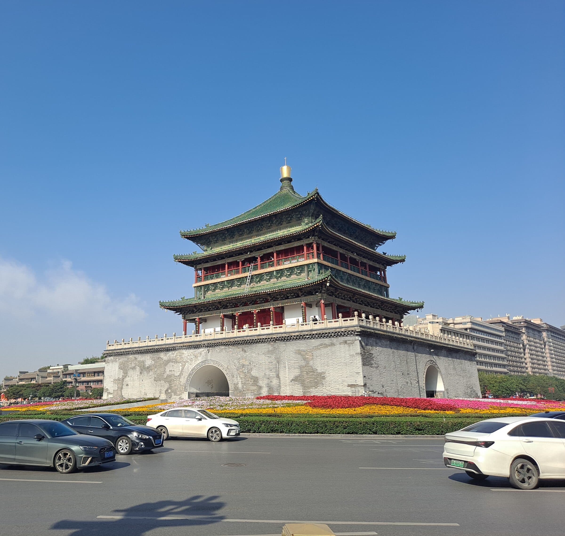 Xi'an's Bell Tower in daylight under a blue sky: a triple-eaved pavilion with green glazed-tile roofs and a golden finial above its brick base and arch, ringed by the flower beds of its traffic roundabout