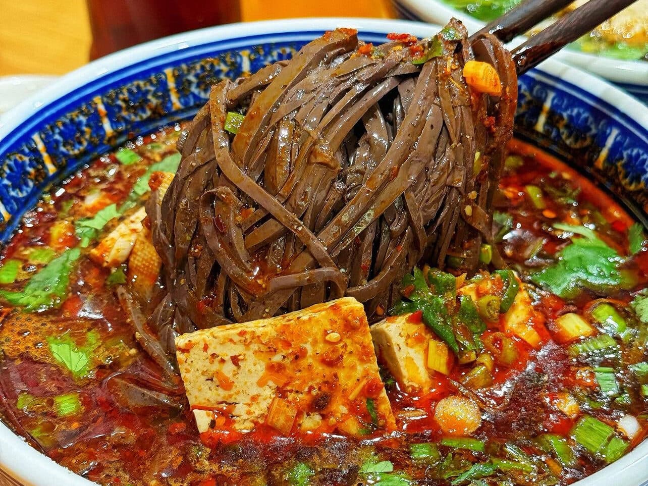 A bowl of dark buckwheat noodles in chilli-oil broth with bread cubes, greens and tofu