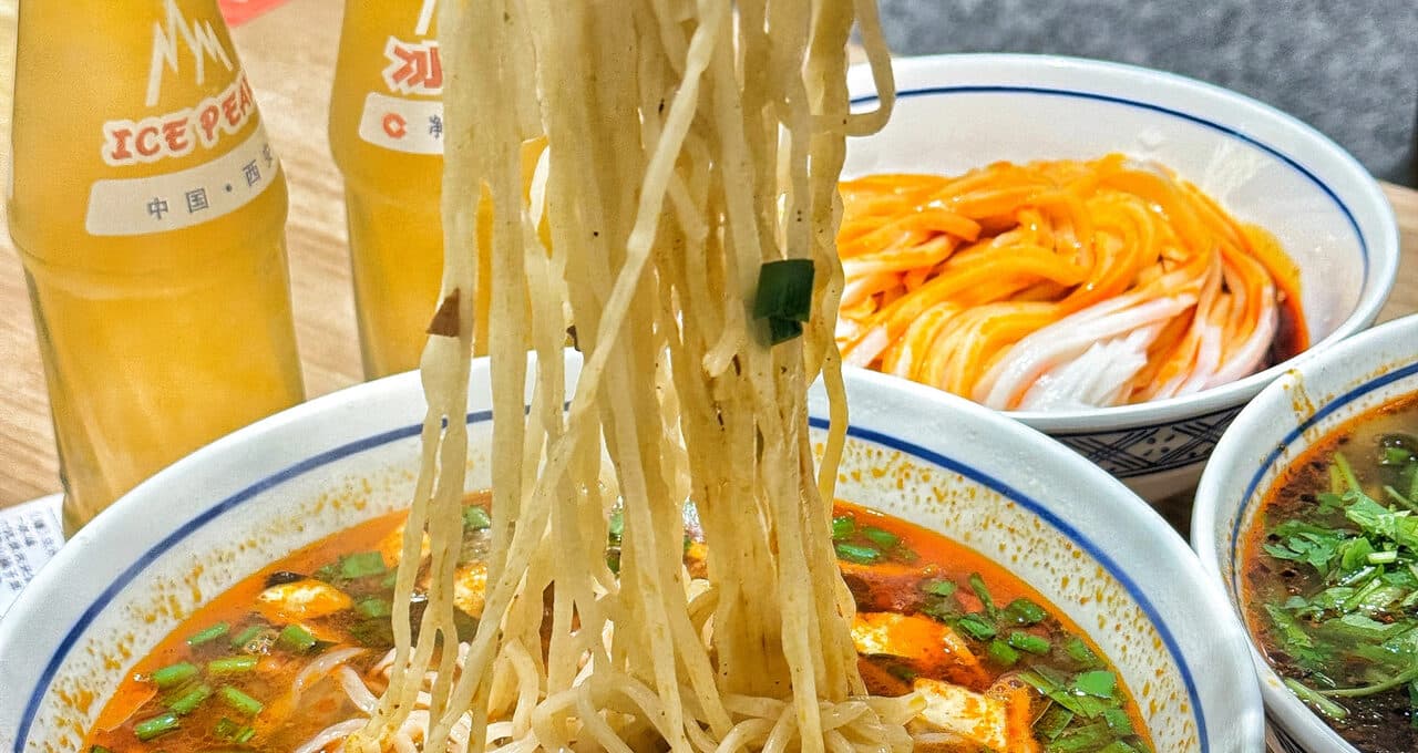 Chopsticks lifting a bundle of noodles from a bowl, with a second bowl of chilli-oil cold noodles and two bottles of Ice Peak soda beside it