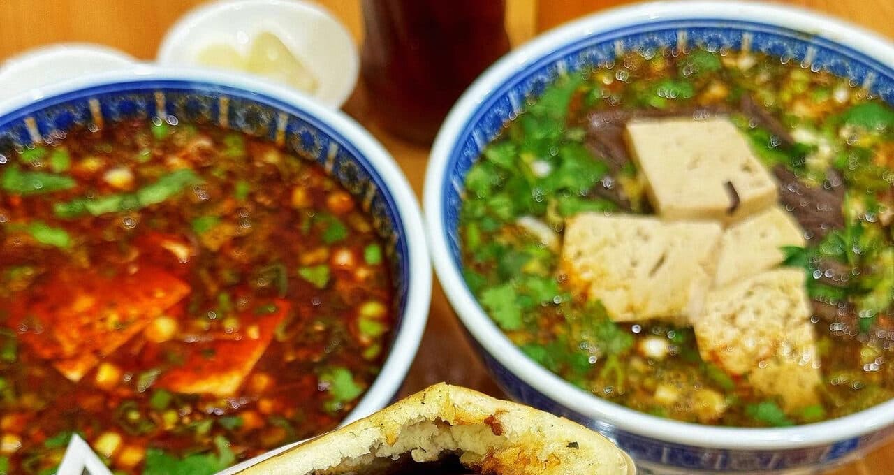 Two bowls of Xi'an paomo: stir-fried paomo in chilli oil at the left and clear-broth paomo at the right, with cubed bread, wood ear and greens in the broth, a torn flatbread and a dish of sweet garlic beside them
