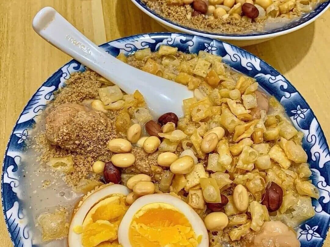 A bowl of Xi'an youcha with fried dough twists: a pale gruel with peanuts, soybeans and half a braised egg, a white porcelain spoon in it