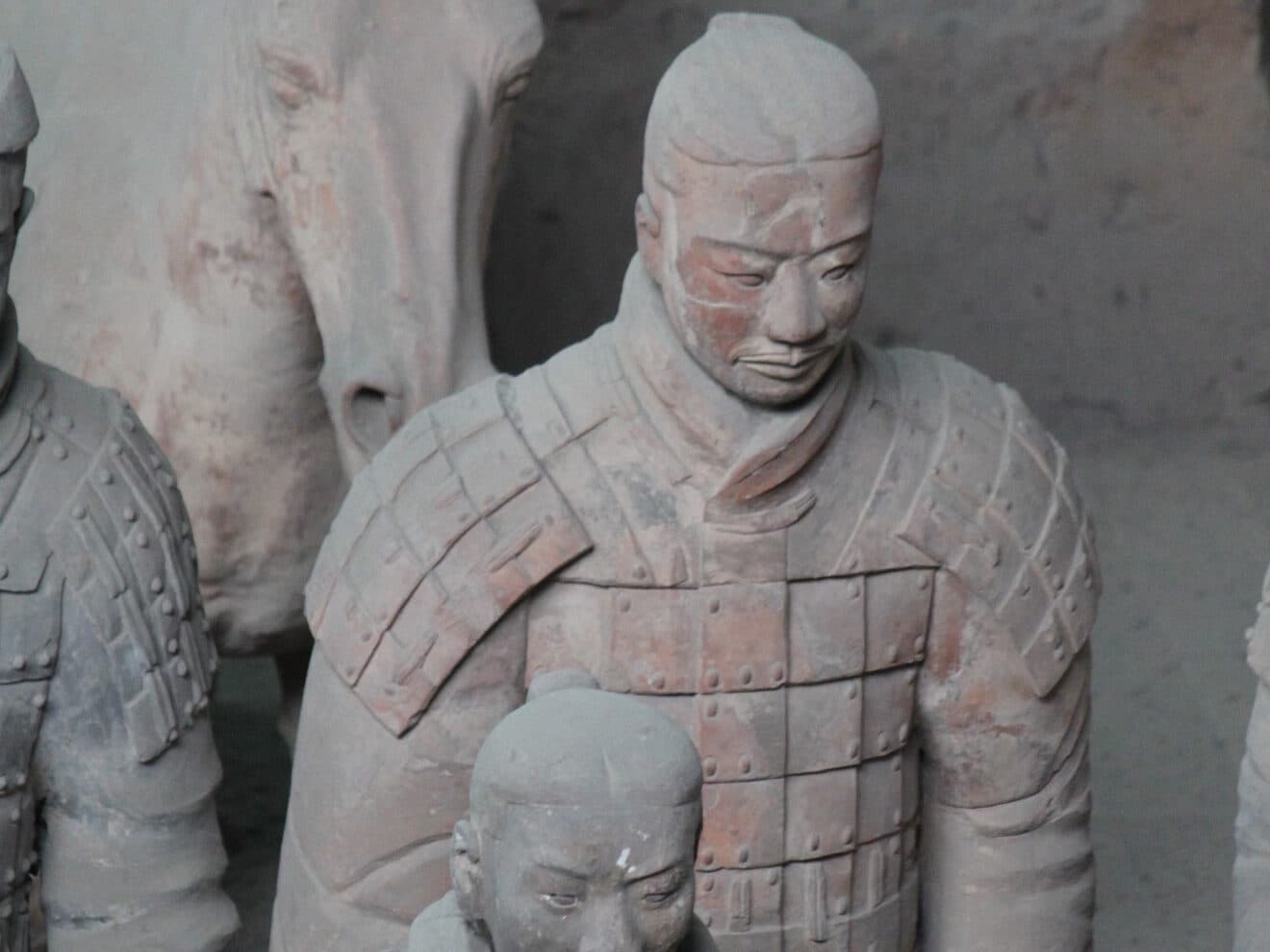 A close view of armoured terracotta warriors in the pit, armour and topknot detail visible, with more figures and a horse's head behind