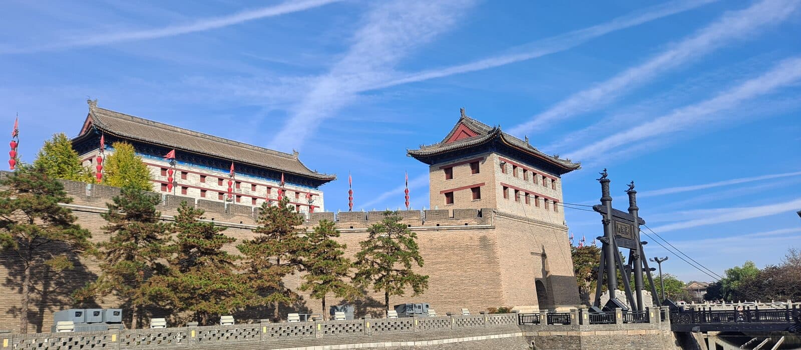 The South Gate (Yongningmen) stretch of Xi'an's city wall in daylight: two towers on the ramparts (a colonnaded gate tower at the left and the brick archer tower with rows of windows at the right), red lanterns and flags along the battlements, the moat-bridge archway at the right under a blue sky with faint contrails
