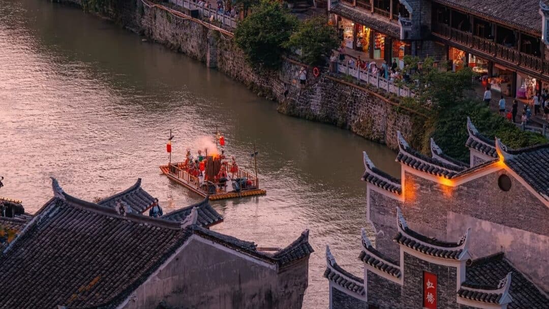 Fenghuang Ancient Town riverside stilt houses and stone embankment at dusk, with a lantern-lit boat on the river