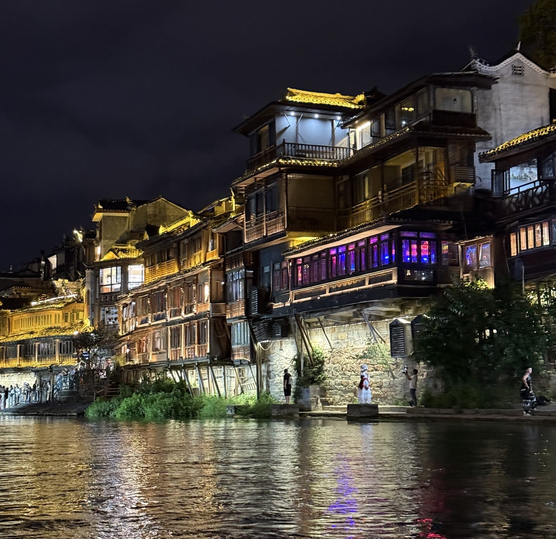 Lit wooden houses on the riverside below the town wall at night