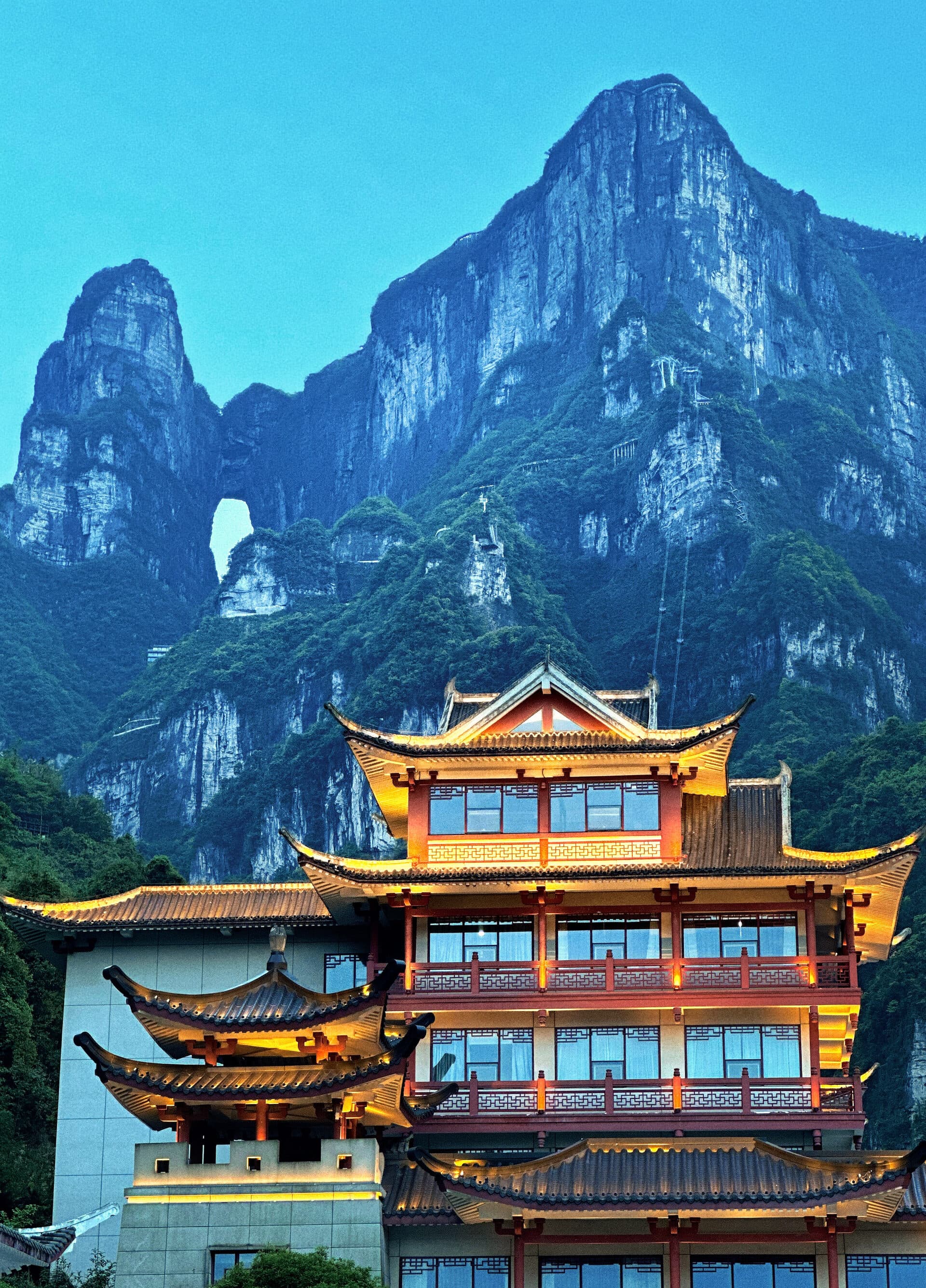 A tiered traditional gate building lit at dusk, with karst peaks and a natural stone arch behind