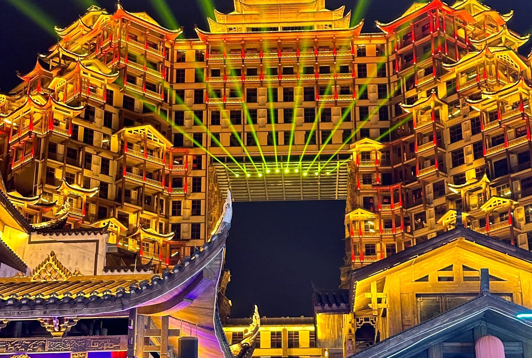 Layered building complex lit gold at night with light beams in the sky