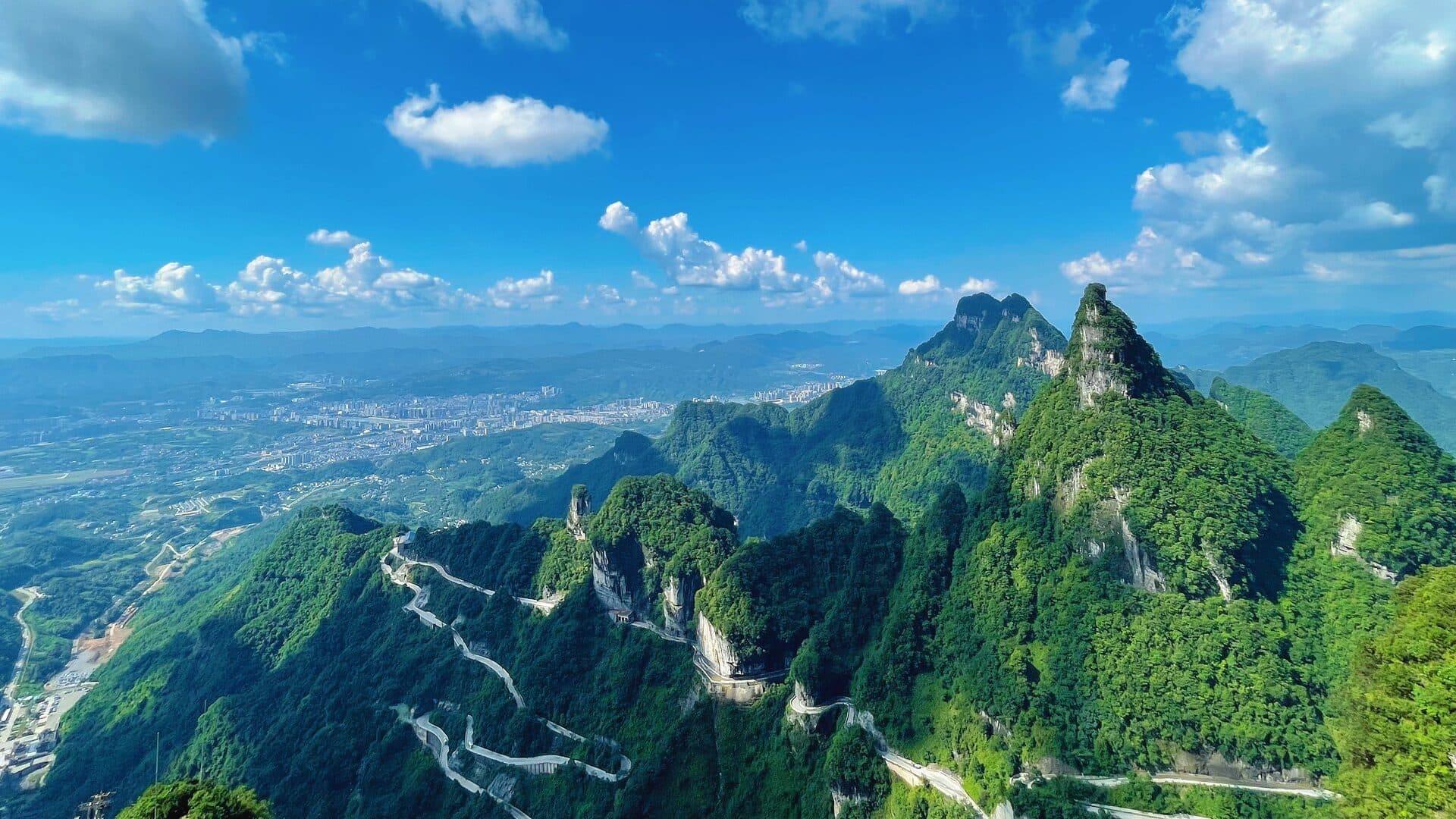 Aerial view of a winding access road through a green karst valley with the city in the distance