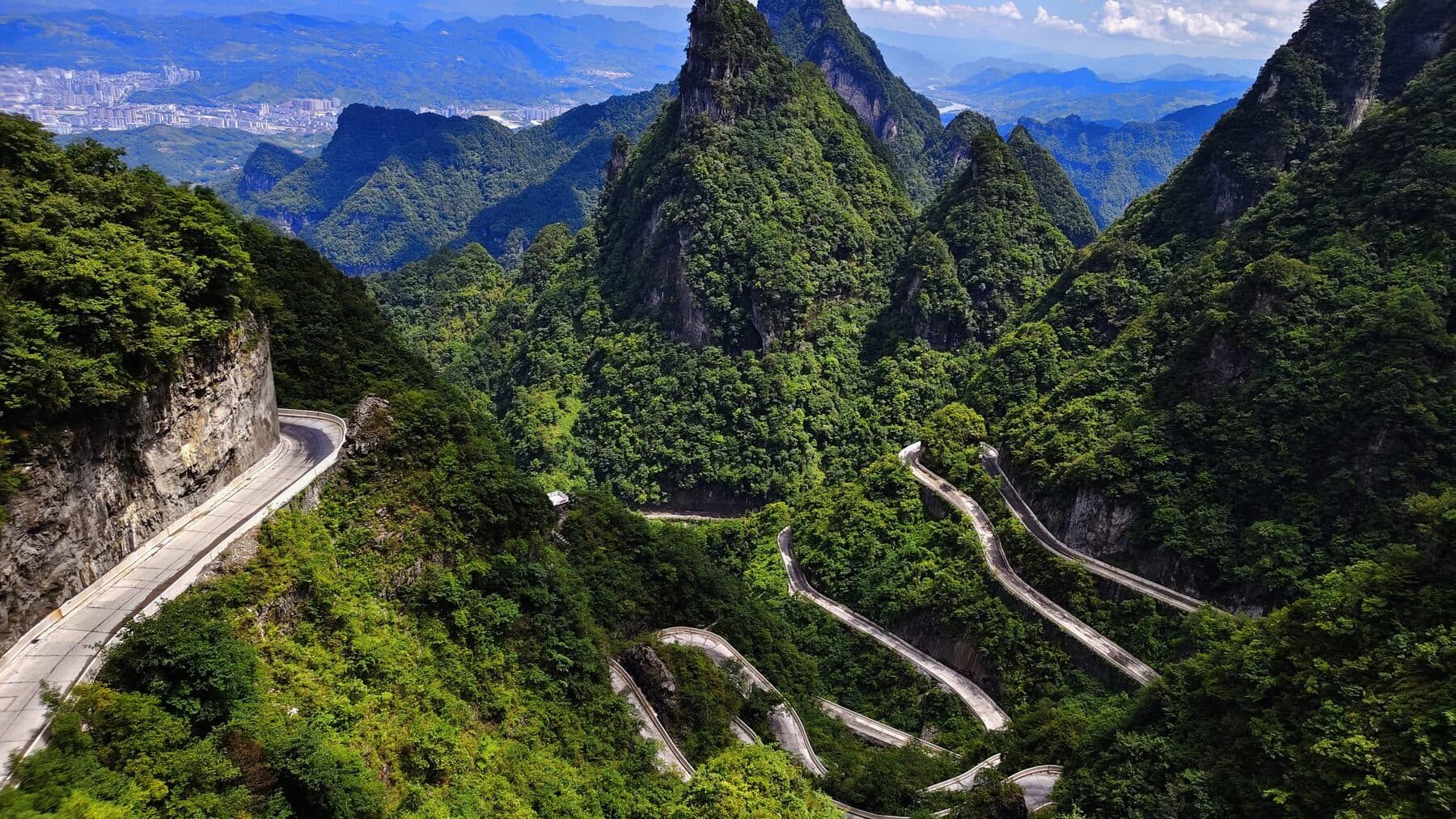 Mountain road switchbacks winding down through green karst peaks toward the city