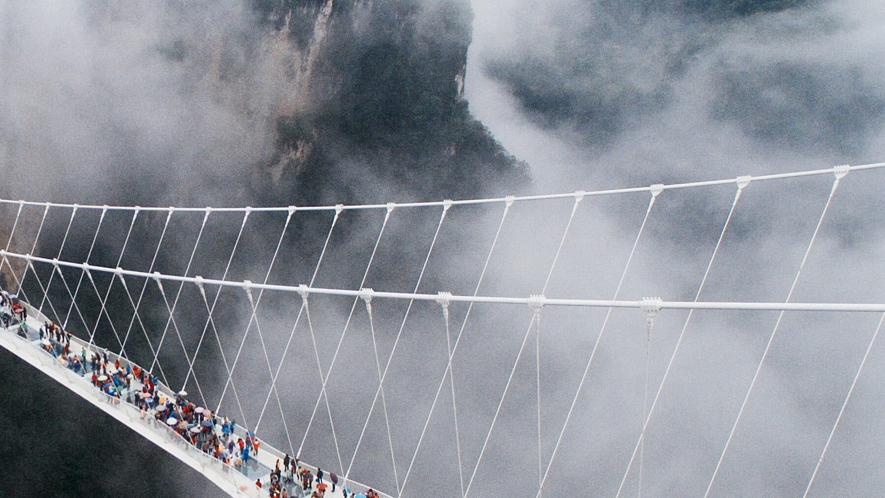 Visitors walking a glass suspension bridge spanning a misty gorge