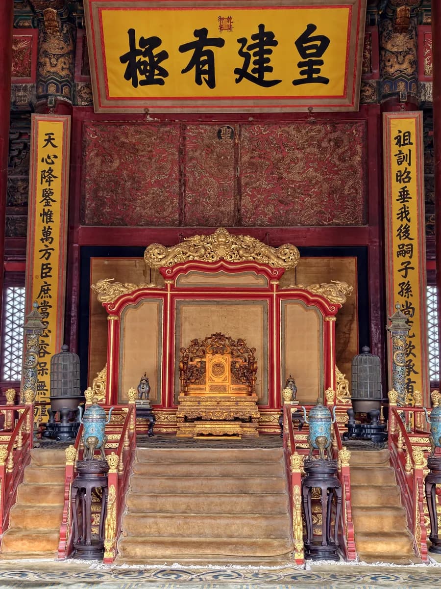 A gilded dragon throne and carved screen inside a palace hall, flanked by lanterns