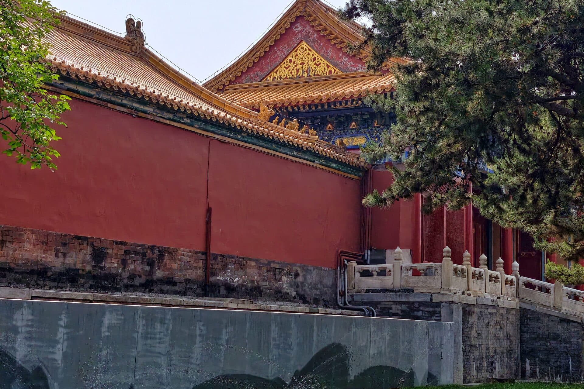 A vermilion palace wall below golden glazed eaves, with a stone balustrade and green trees